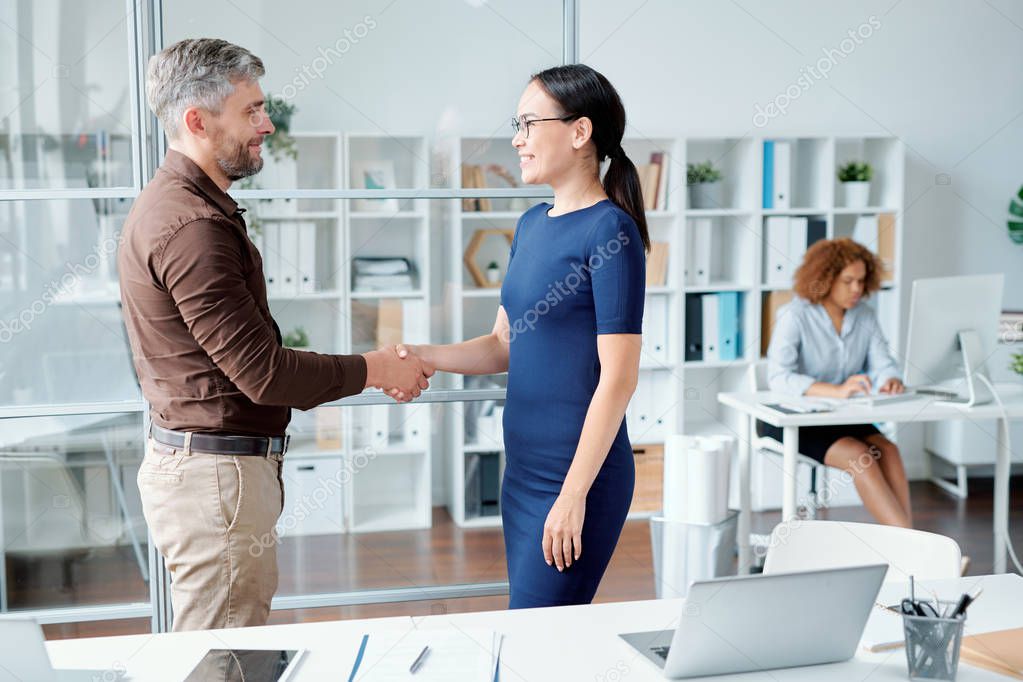 Young successful colleagues or business partners in smart casual shaking hands by workplace after making agreement about working points