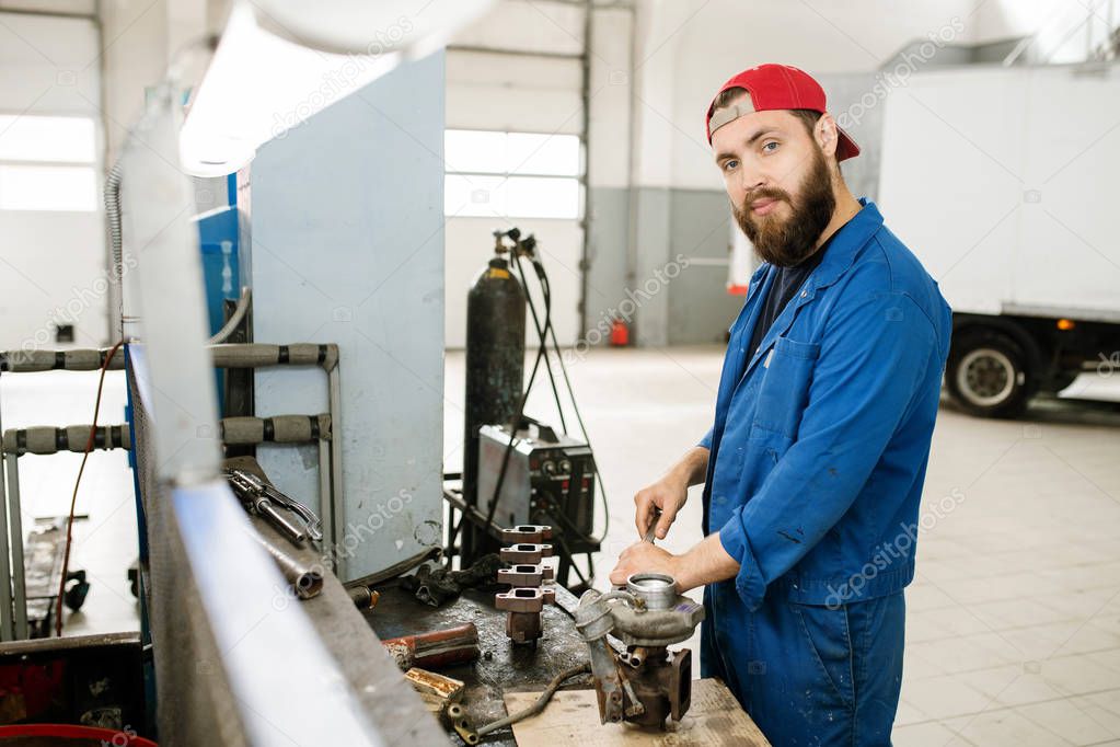 Young bearded repairman in workwear looking at you while using wrench or other handtool to fix parts of car engine