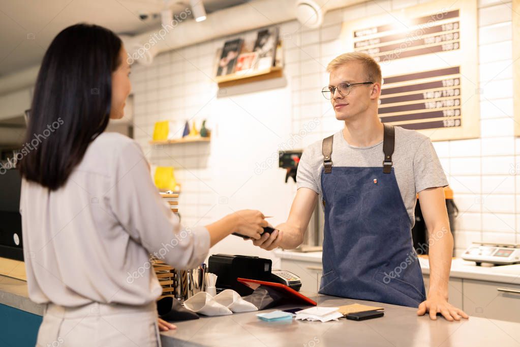 Young waiter holding electronic payment machine over counter while one of clients paying by credit card for her order