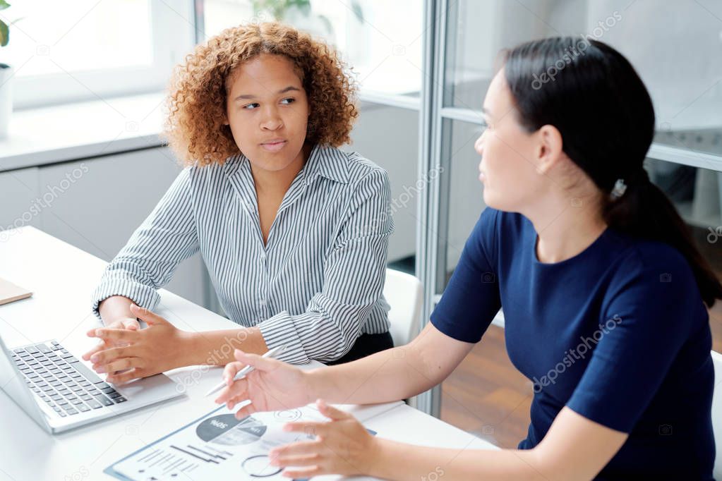 Young serious mixed-race accountant looking at her colleague explaining details of financial development of their company