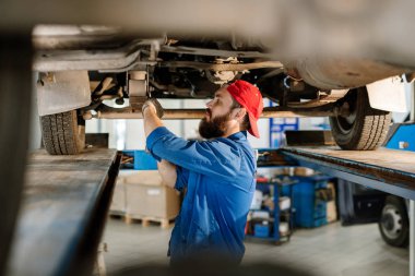 İş kıyafetli genç adam arabanın altında duruyor ve bir arabanın bakımını yaparken ya da tamir ederken iş okulunu kullanıyor.