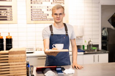 Young handsome blond waiter in eyeglasses and workwear giving you tray with cup of coffee while standing by counter clipart