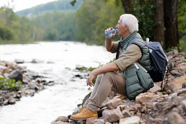 Trött Mogen Backpacker Dricksvatten Från Plastflaska Sittande Flodstranden Naturlig Miljö — Stockfoto