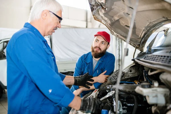 Young bearded worker of technical service looking at his mature colleague examining broken engine of large machine while listening to him