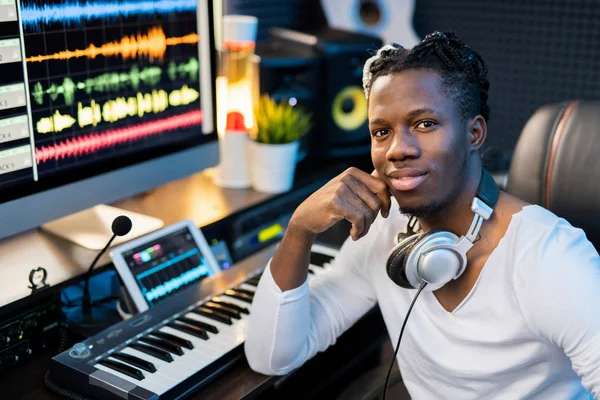快乐的年轻 面带微笑 头戴耳机的混血男子坐在工作场所 手持钢琴键盘和电脑显示器 — 图库照片