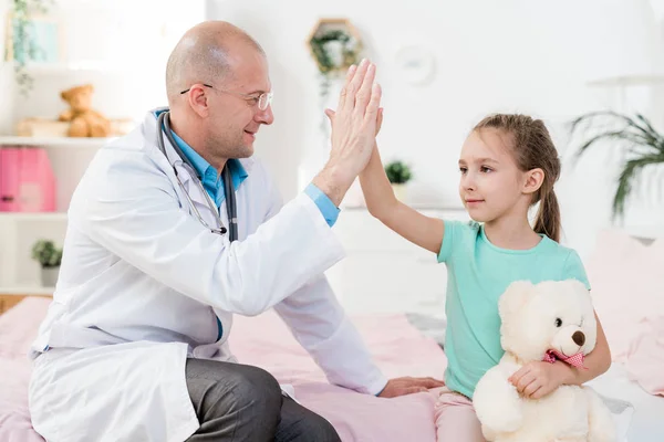 带着玩具熊的可爱小女孩和她的医生坐在医院里互相击掌 — 图库照片