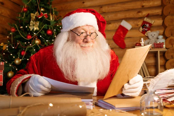 圣诞老人惊奇地坐在桌子旁边 一边看着手里的信封 一边想把信放进信封里 — 图库照片