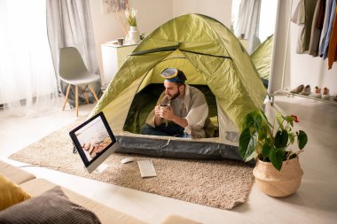 Kafasında dalış maskesi olan ciddi genç sakallı bir adam evdeki çadırda oturmuş bilgisayar ekranında deniz fotoğrafına bakarken çay içiyor.