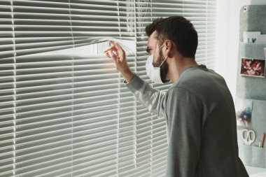 Tıbbi maskeli ve kazaklı sakallı hasta bir adam. Pencere kenarında duruyor ve hastalık sırasında evde otururken külüstürden dışarı bakıyor.