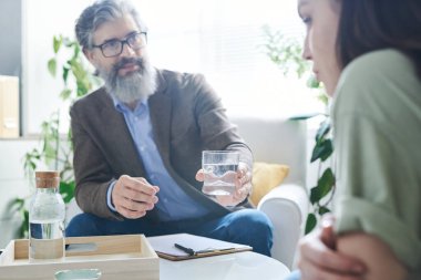 Ofisteki masada konuşurken genç bayan hastasına sakallı bir bardak su uzatan kıdemli danışman.