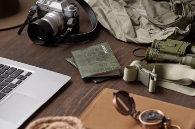 Yeşil kapaklı pasaportların, kemerin, dürbünün, fotoğraf makinesinin, ceketin ve laptopun ahşap masadaki yakın plan görüntüleri.