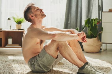 Spor ayakkabılı ve şortlu genç sporcu evde sıkı çalıştıktan sonra gözleri kapalı halıya oturmuş dinleniyor.