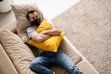 Yüzü maskeli genç bir adamın Coronavirus izolasyonu sırasında yastığı kucaklaması ve kanepede uyuması.