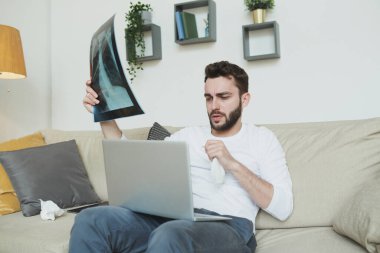 Mendili ve akciğeri olan sakallı genç bir adam dizüstü bilgisayarın önündeki koltukta oturuyor ve evdeki online doktorla görüşüyor.