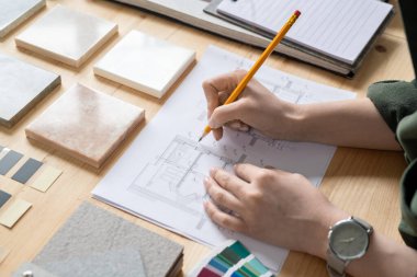 Masanın başında otururken, kağıt çizim planı üzerine çizim yapan iç mimarın elleri.