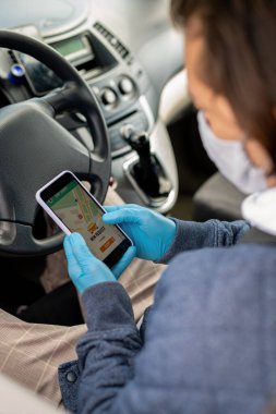 Koruyucu eldivenli ve maskeli çağdaş taksi şoförü direksiyonda oturuyor ve sürmeden önce ekranda navigatör uygulaması ile akıllı telefon kullanıyor.