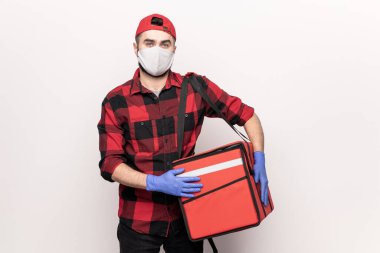 Kapşonlu erkek kurye, kırmızı flanel, koruyucu maske ve eldiven karantina döneminde çalışırken kahve müşterilerinin siparişleriyle dolu büyük bir çanta taşıyor.