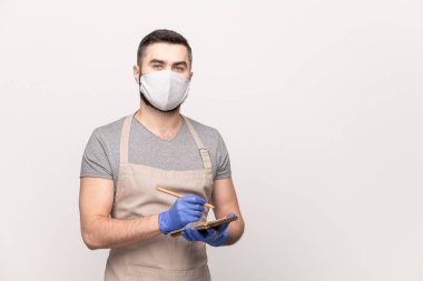 Önlüklü, koruyucu maskeli ve eldivenli genç garson kafede ya da restoranda çalışırken siparişinizi not defterine yazıyor.