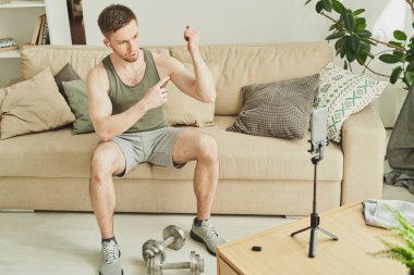 Aktif genç kaslı adam, akıllı telefon kamerasının önünde formda kalmak için ne yaptığını açıklarken pazılarını işaret ediyor.