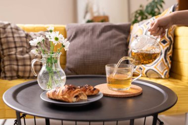 Sabah kahvaltıda kruvasan yerken çaydanlığa yeşil bitki çayı döken genç bir kadının eli.