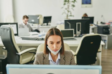 Karantina sırasında yarı boş bir ofiste çalışırken modern bilgisayar kullanarak çenesinde maske olan çekici genç bir kadın.