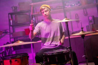 Tişörtlü, kot pantolonlu ve bereli bir adam garajda davul setinin önünde oturuyor ve prova sırasında davul ve davula vuruyor.