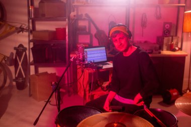 Genç, ciddi bir müzisyen garajda davul seti başında dizüstü bilgisayar, müzik aletleri ve çalışma aletleriyle sana bakıyor.