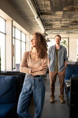 Bej renkli kazaklı ve mavi kotlu mutlu bir kadın kanepede dikilip kafenin penceresinden bakarken kollarını kavuşturuyor.