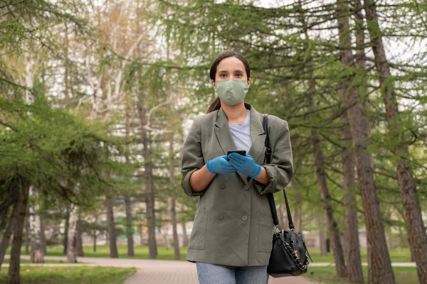 Young brunette woman in protective gloves and mask looking at you while using mobile phone on her way home along public park
