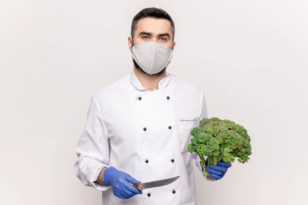 Young male chef in protective mask, gloves and white uniform holding sharp knife and fresh broccoli while going to cook vegetable soup