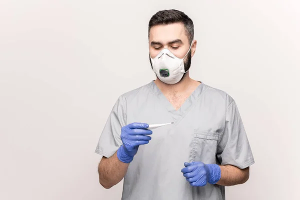 病気の患者の温度を測定しながら 灰色の均一で青い手袋と温度計を見て白いマスクの若い医師 — ストック写真