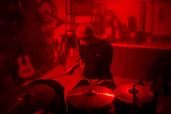 Contemporary musician in casualwear recording his music while sitting in front of drumset in red lit room or garage and hitting drums