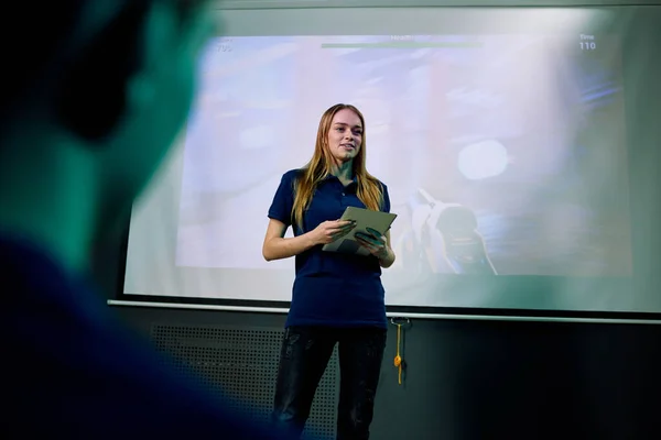 新型网络游戏的年轻而自信的女性开发者 带数字平板电脑 在演示期间站在大屏幕旁边 — 图库照片