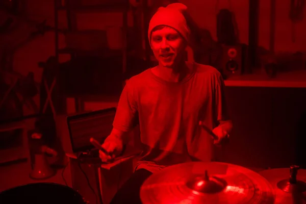 Young musician in beanie and t-shirt hitting cymbals and drums with drumsticks while sitting by drum kit and laptop in garage