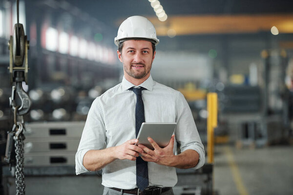 Portrait of confident handsome technical engineer in hardhat using tablet in industrial shop of large plant