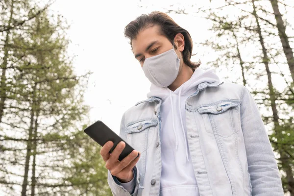 재킷을 후드를 마스크를 스마트폰을 메시지를 보내는 젊은이가 여름날 공원에서 산책을 — 스톡 사진