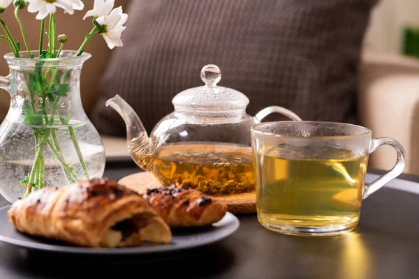 緑茶のティーポットとカップ 皿の上の新鮮なクロワッサン 肘掛け椅子の枕を背景にテーブルの上の花の束 — ストック写真