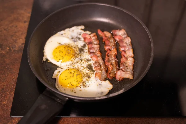 台所で朝食のための料理を調理する過程で電気ストーブの上で揚げ卵とベーコンとフライパン — ストック写真
