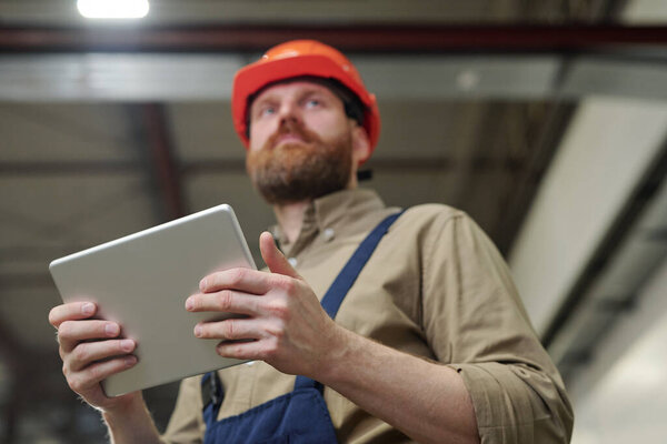 Below view of young bearded engineer in hardhat using tablet while controlling work at factory