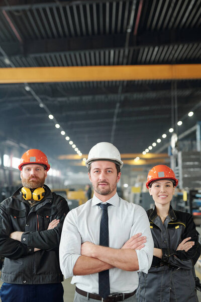 Confident factory team of professionals in hardhats standing with crossed arms in contemporary shop of large plant