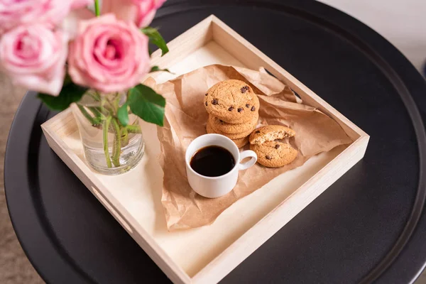 紙にコーヒーとクリスピークッキーのカップと低正方形のカートンボックス 小さな黒丸テーブルの上にガラスのピンクのバラの束 — ストック写真