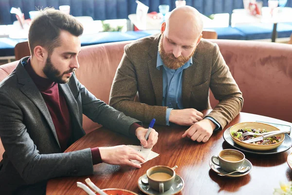 有思想的年轻同事们坐在桌旁 一边吃着沙拉和咖啡杯 一边在餐巾上写下自己的商业理念 — 图库照片