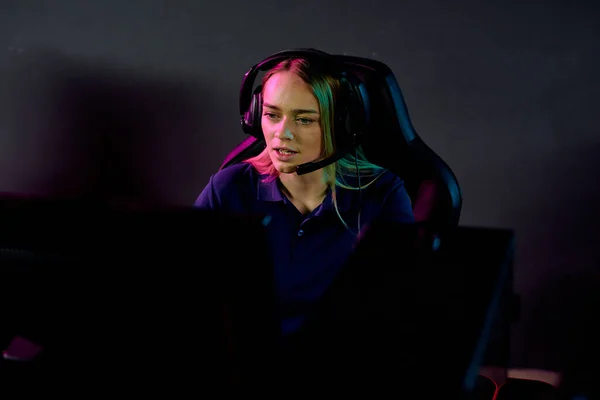 年轻而严肃的金发女电子体育游戏玩家坐在监控器前的黑暗俱乐部里 头戴耳机 看着电脑屏幕 — 图库照片