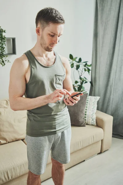 Happy young man in sportswear scrolling in smartphone while searching for online video course of home fitness while standing in living-room