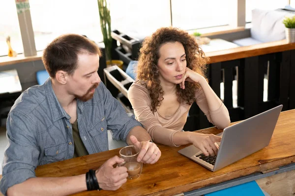 当代的一对年轻夫妇坐在自家咖啡店的木桌旁 卷曲的头发女人们指着笔记本电脑的显示屏 — 图库照片