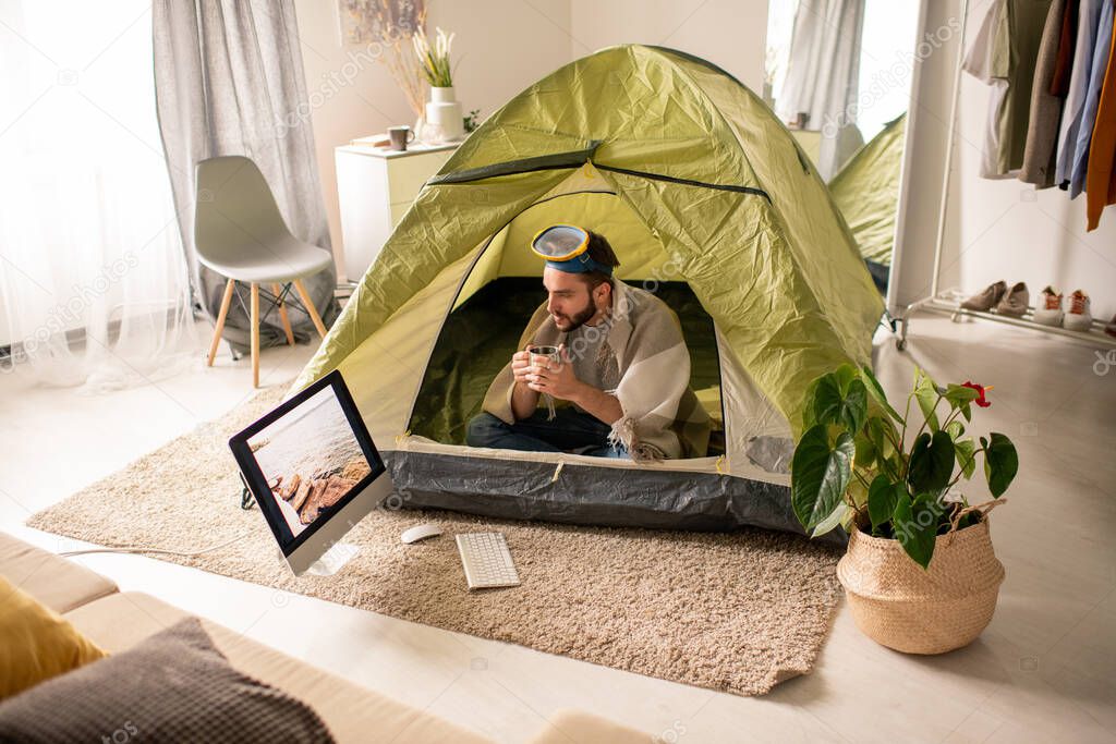 Serious young bearded man with diving mask on head sitting in tent at home and drinking tea while looking at sea picture on computer screen
