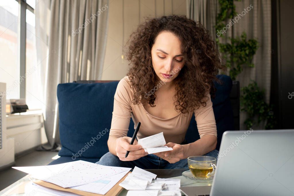 Young serious businesswoman with long wavy hair looking through payment bills while sitting on couch in front of laptop