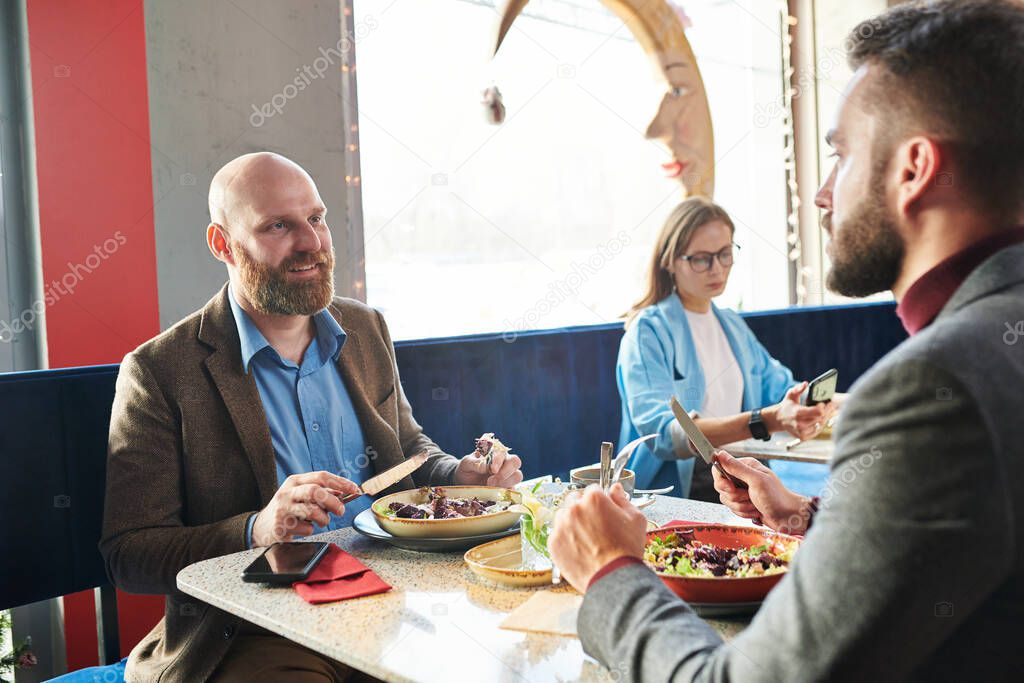 Smiling bald businessman sitting at table and eating salad while chatting with colleague over lunch in cafe