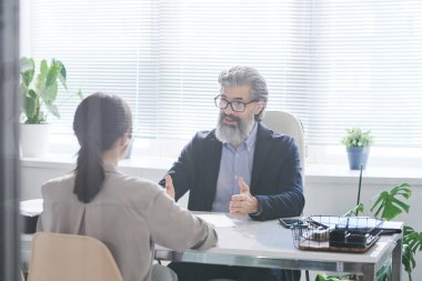 Gri sakallı, kendinden emin bir işveren ofisinde konuşurken genç başvuru sahibine bir şey anlatıyor.