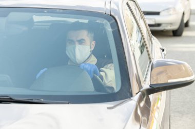 Koruyucu maskeli ve eldivenli genç bir kurye karantina sırasında müşterilerine mal teslim ederken arabasında oturuyor.